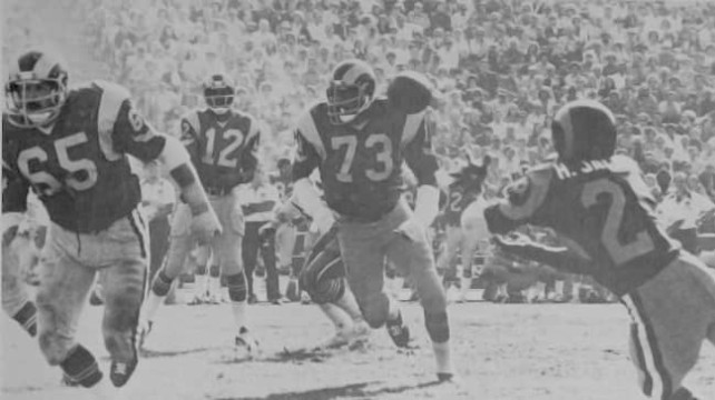 In the mid-1970s the Rams possessed one of the best offenses in the NFL. Here we see quarterback James Harris (#12), a Pro Bowler in 1974, connecting with All Pro receiver Harold Jackson (#29) as Pro Bowl lineman Tom Mack (#65) and Charlie Cowan (#73) pull out to lead.  