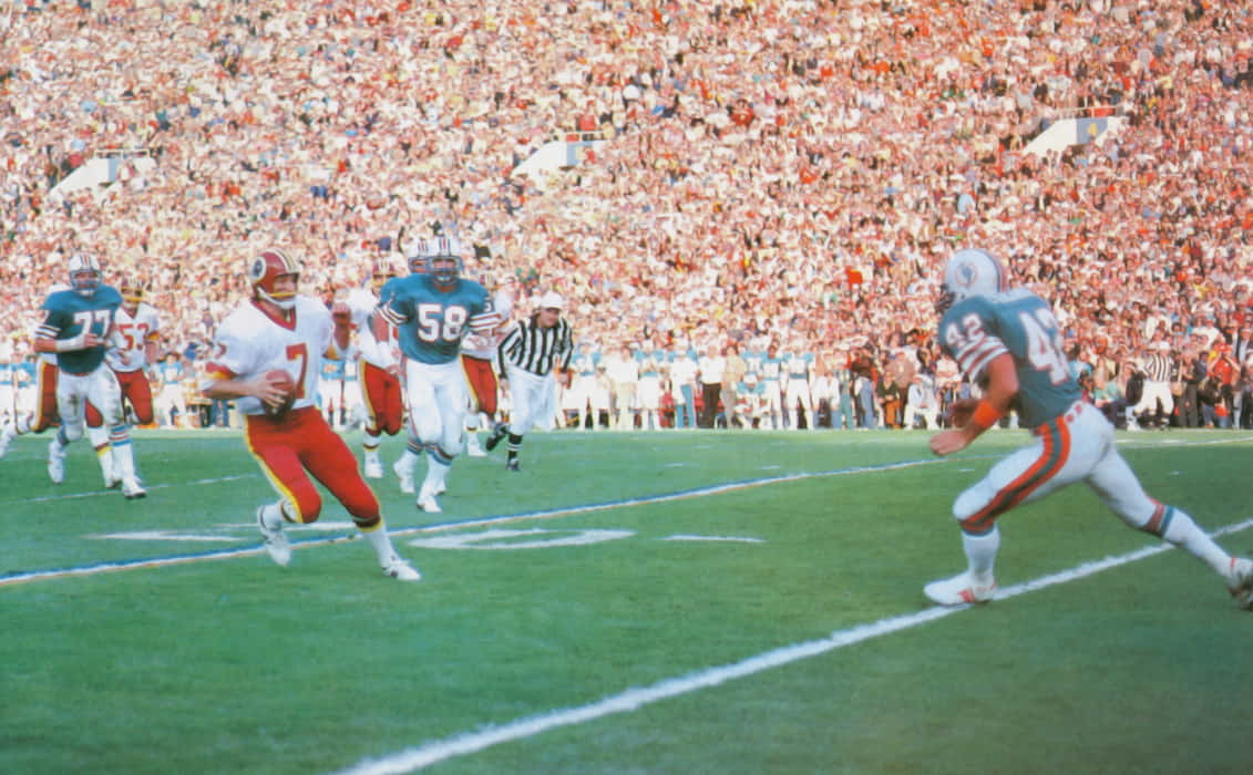 In Super Bowl XVII, Joe Theismann gains yardage as spectators watch, with Lyle Blackwood closing in to make a tackle.