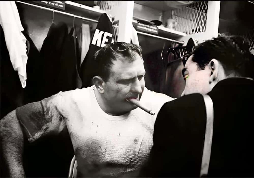 Black and white photo of Packers guard Fuzzy Thurston smoking a cigar in the locker room.