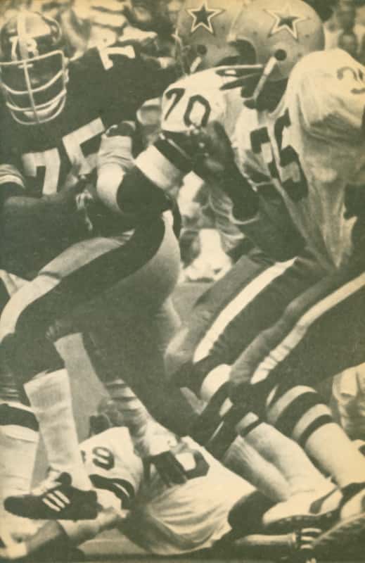 A black and white photo capturing Calvin Hill advancing the ball against the Steelers, with Rayfield Wright blocking Mean Joe Greene.